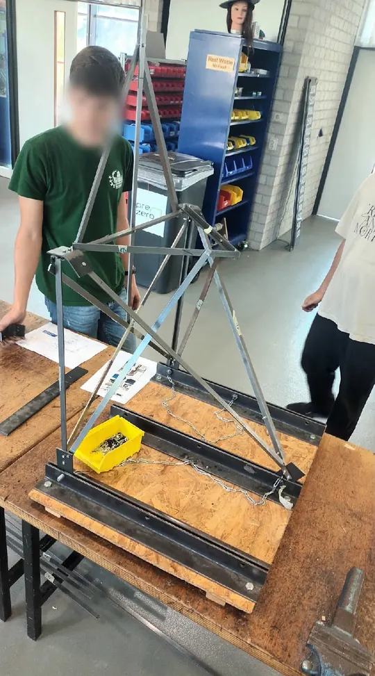 A steel truss structure on a wooden base in the workshop, with a load hanging from it.